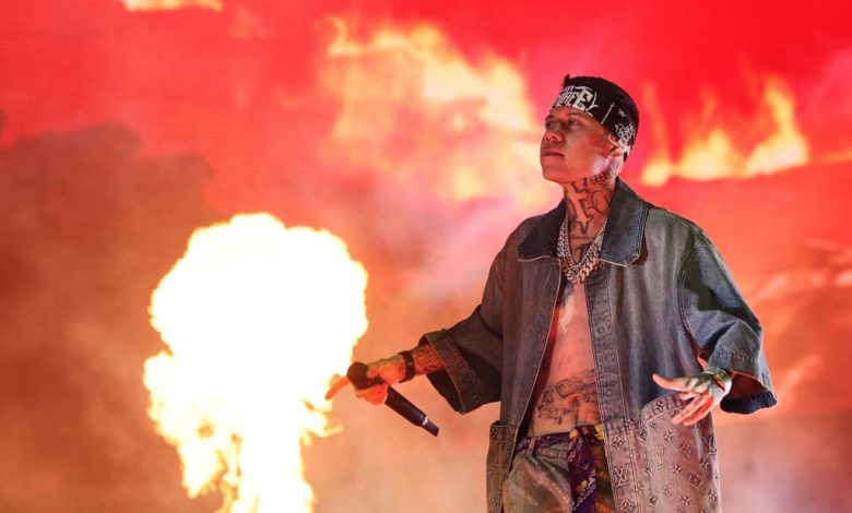 Santa Fe Klan, a tattooed rapper, performs on stage with a microphone, wearing a denim jacket and headscarf, against a backdrop of dramatic red flames and smoke at Coachella.
