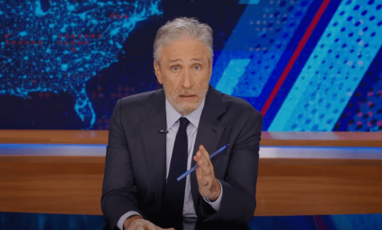 A mature man with graying hair and a beard sits behind a desk in a television studio, speaking and gesturing with a pen. He wears a dark suit and tie, with a blue and red