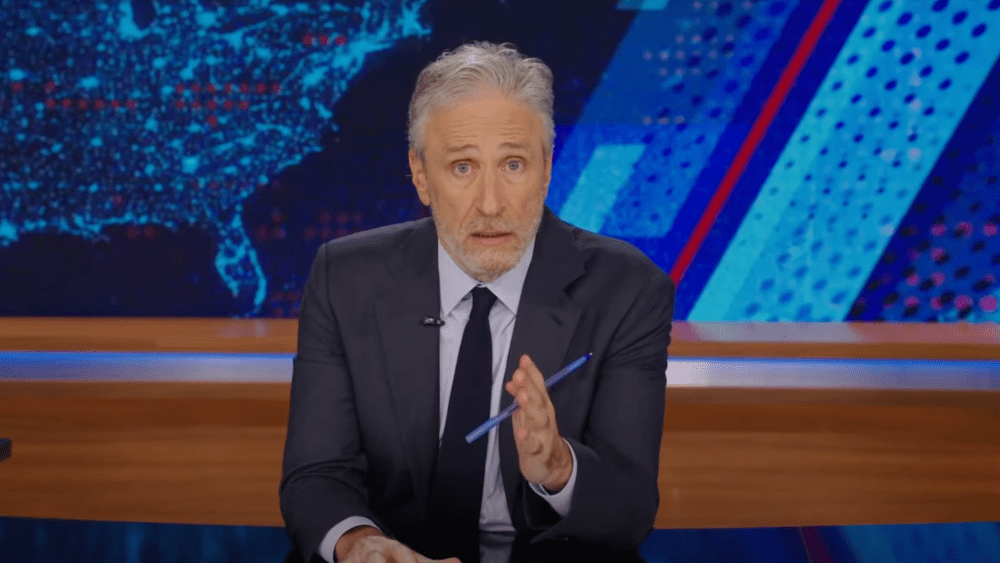 A mature man with graying hair and a beard sits behind a desk in a television studio, speaking and gesturing with a pen. He wears a dark suit and tie, with a blue and red