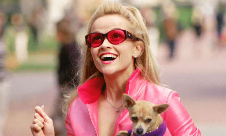 A joyous woman with blonde hair, reminiscent of Reese Witherspoon, wears large red sunglasses and a bright pink jacket, holding a small chihuahua in her arms, both smiling and walking