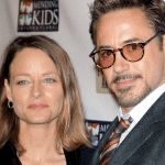 Two adults at a formal event, one wearing a gray suit and round glasses, the other in a dark jacket, standing in front of a backdrop with charity logos related to addiction awareness.