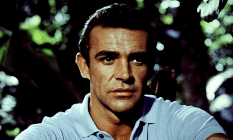 A close-up of a serious-looking middle-aged man with dark, combed-back hair, reminiscent of James Bond. He's wearing a light blue polo shirt, set against a backdrop of dense green foliage