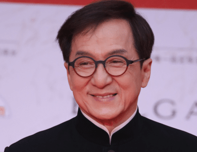A middle-aged Asian man in formal attire smiles gently. He wears a black traditional shirt with a high collar and glasses. The background features a red event banner with white text and logos mentioning Jackie Chan Ass