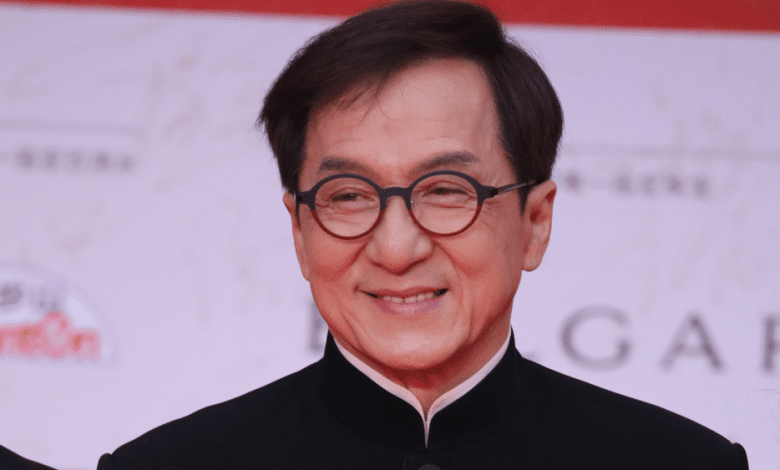 A middle-aged Asian man in formal attire smiles gently. He wears a black traditional shirt with a high collar and glasses. The background features a red event banner with white text and logos mentioning Jackie Chan Ass