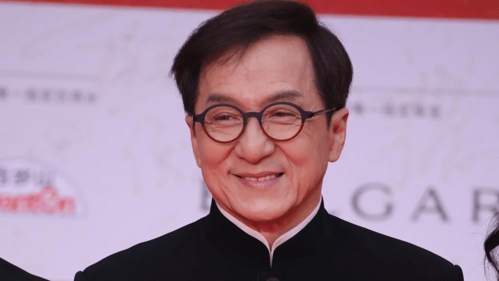 A middle-aged Asian man in formal attire smiles gently. He wears a black traditional shirt with a high collar and glasses. The background features a red event banner with white text and logos mentioning Jackie Chan Ass