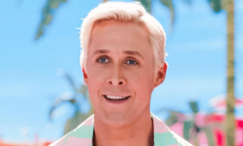 A man with short blond hair and fair skin smiles at the camera. He's wearing a pastel-colored shirt with a tropical design featuring iconic images of Gosling. The background shows a bright blue sky