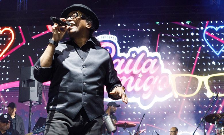 A male singer in a black suit and hat performs on stage with a microphone, backed by musicians and vibrant lights at the Baila Conmigo Fest. A large sign saying "baile night