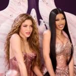 Two women in glamorous dresses sit side by side with smiles, against a large, decorative pink winged backdrop adorned with crystals. One woman has long, wavy blonde hair, and the other has straight