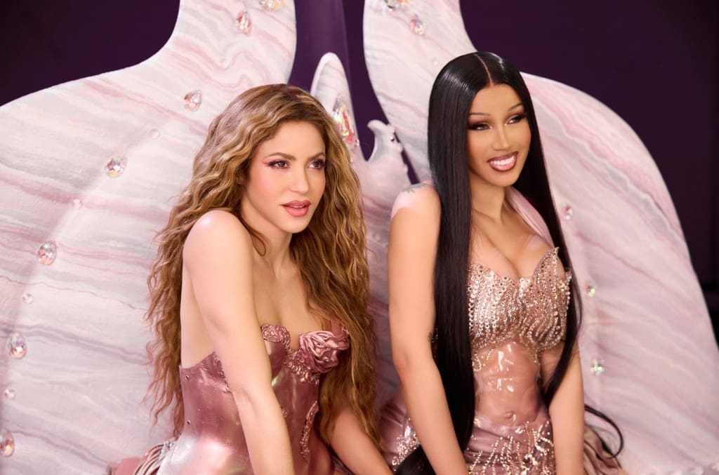 Two women in glamorous dresses sit side by side with smiles, against a large, decorative pink winged backdrop adorned with crystals. One woman has long, wavy blonde hair, and the other has straight
