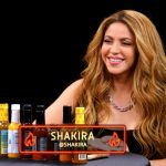 Shakira is seated at a table smiling, with a variety of colorful hot sauce bottles lined up in front of her on the 'Hot Ones' talk show. A screen with her name and a