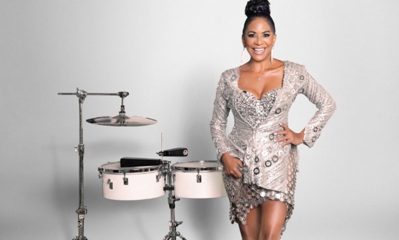 A woman with an elegant updo hairstyle, wearing a glittery silver dress, smiling and standing confidently in front of a set of silver drums on a gray background discusses her salsa debut.