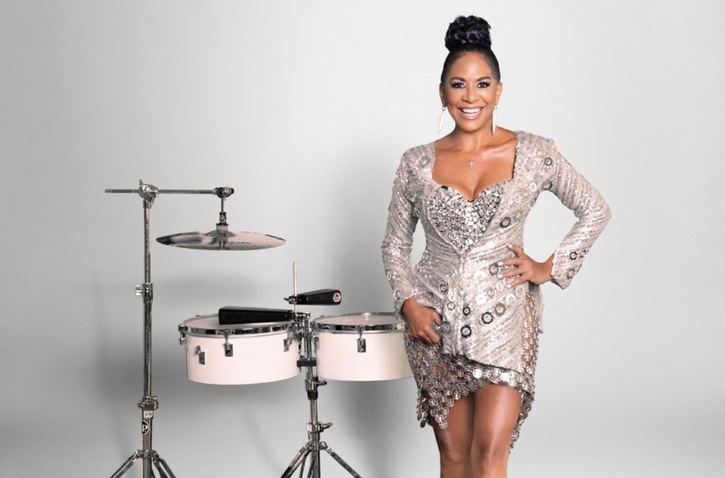 A woman with an elegant updo hairstyle, wearing a glittery silver dress, smiling and standing confidently in front of a set of silver drums on a gray background discusses her salsa debut.