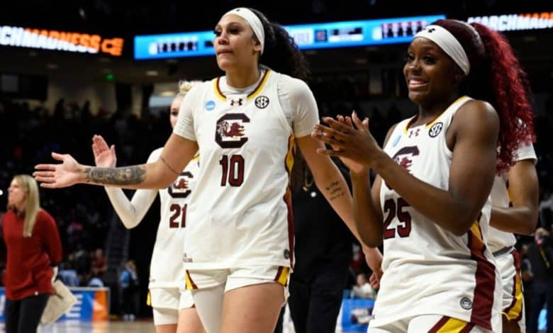 Two female basketball players from South Carolina, wearing white jerseys numbered 10 and 25, joyfully walk along the court, clapping hands during a game. The crowd and a banner reading "202