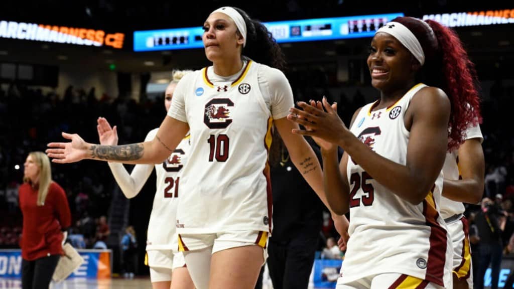 Two female basketball players from South Carolina, wearing white jerseys numbered 10 and 25, joyfully walk along the court, clapping hands during a game. The crowd and a banner reading "202
