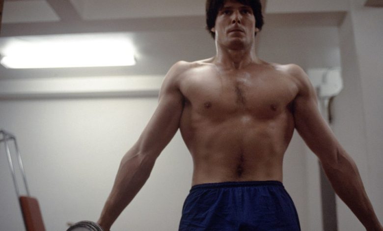 A shirtless man with a toned physique stands in a dimly lit room, gripping a pull-up bar with a focused expression, wearing blue shorts reminiscent of Christopher Reeve's Superman costume. His defined