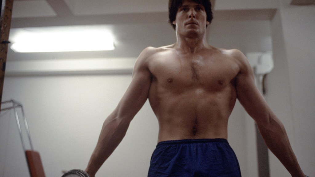 A shirtless man with a toned physique stands in a dimly lit room, gripping a pull-up bar with a focused expression, wearing blue shorts reminiscent of Christopher Reeve's Superman costume. His defined