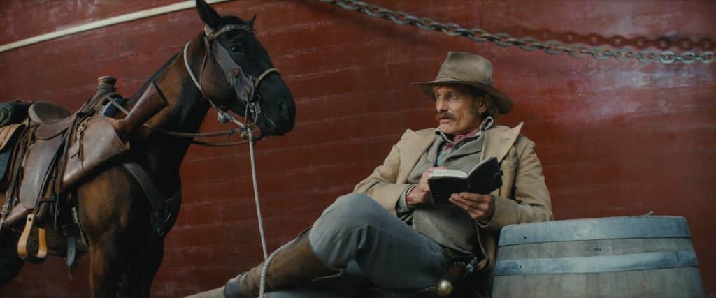A man dressed in vintage cowboy attire sits beside a black horse, holding an open book and a pipe. He’s resting against a large red metal structure from Viggo Mortensen's 'The