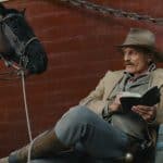 A man dressed in vintage cowboy attire sits beside a black horse, holding an open book and a pipe. He’s resting against a large red metal structure from Viggo Mortensen's 'The