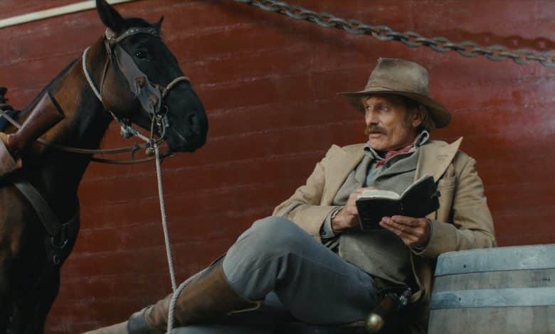 A man dressed in vintage cowboy attire sits beside a black horse, holding an open book and a pipe. He’s resting against a large red metal structure from Viggo Mortensen's 'The
