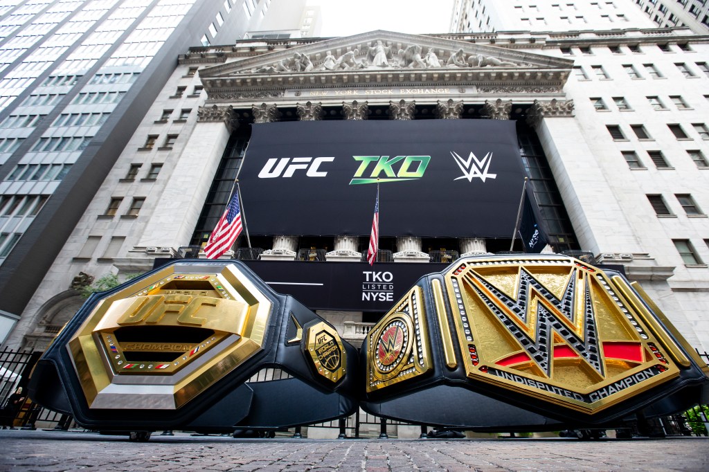 Two championship belts, one labeled "ufc" and the other "wwe," are displayed in front of a large screen showing ufc tko and wwe logos, with the New York Stock
