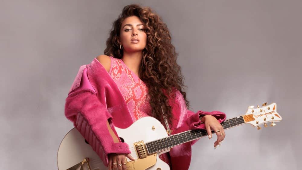 A woman with voluminous curly hair, reminiscent of Tori Kelly's style, holds a white electric guitar, wearing a vibrant pink lace dress and a matching oversized blazer, against a soft gray background