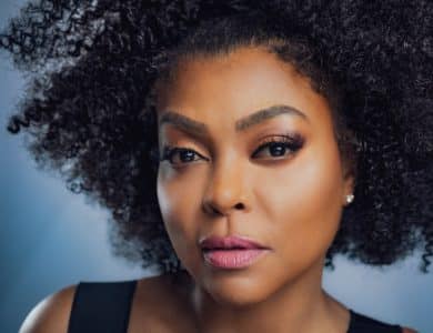 Close-up portrait of a black woman with voluminous curly hair, flawless makeup, and a serious expression. She looks directly at the camera, her eyebrows slightly furrowed. The background is soft and