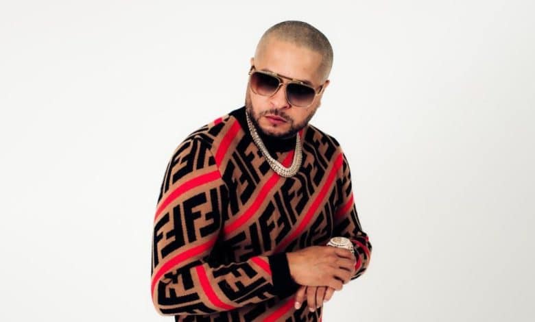 A man in a patterned sweater and sunglasses looks confidently at the camera, with one hand adjusting his watch and a chain necklace visible. The background is plain white, highlighting his stance and attire as Tempo