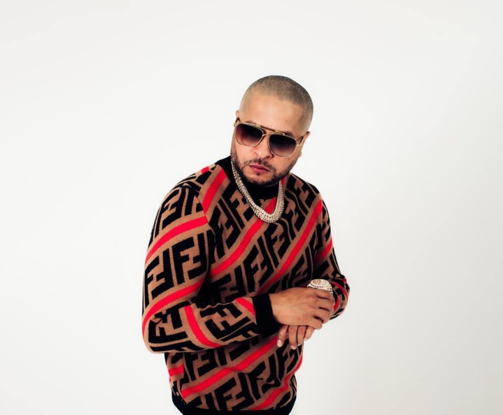 A man in a patterned sweater and sunglasses looks confidently at the camera, with one hand adjusting his watch and a chain necklace visible. The background is plain white, highlighting his stance and attire as Tempo