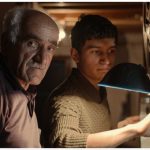 An older man with gray hair and a younger man focus on a vintage film projector in a dimly lit room, suggesting a moment of teaching or shared interest in the "Return of the Projectionist