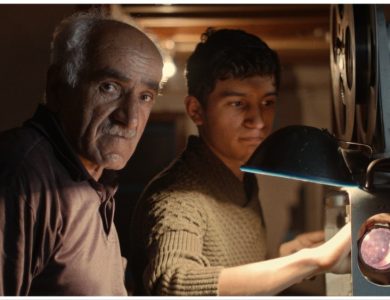 An older man with gray hair and a younger man focus on a vintage film projector in a dimly lit room, suggesting a moment of teaching or shared interest in the "Return of the Projectionist