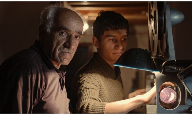 An older man with gray hair and a younger man focus on a vintage film projector in a dimly lit room, suggesting a moment of teaching or shared interest in the "Return of the Projectionist