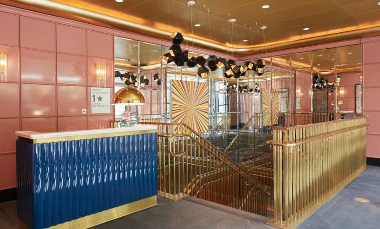 A luxurious hotel lobby featuring a blue reception desk with gold accents in front. The area has pink paneled walls, mirrored columns, bold geometric floor patterns, and contemporary hanging lights. A staircase with gold