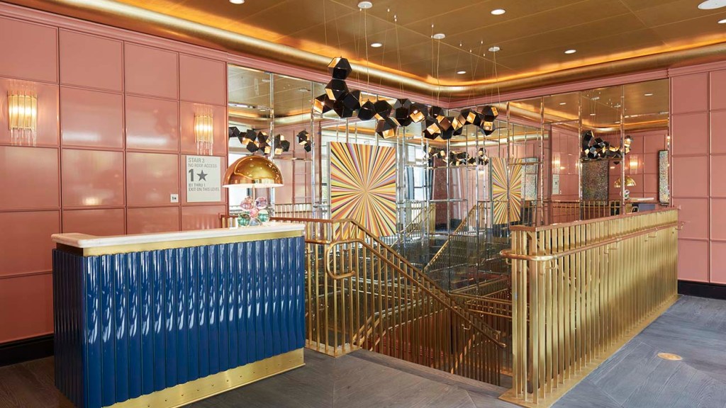A luxurious hotel lobby featuring a blue reception desk with gold accents in front. The area has pink paneled walls, mirrored columns, bold geometric floor patterns, and contemporary hanging lights. A staircase with gold