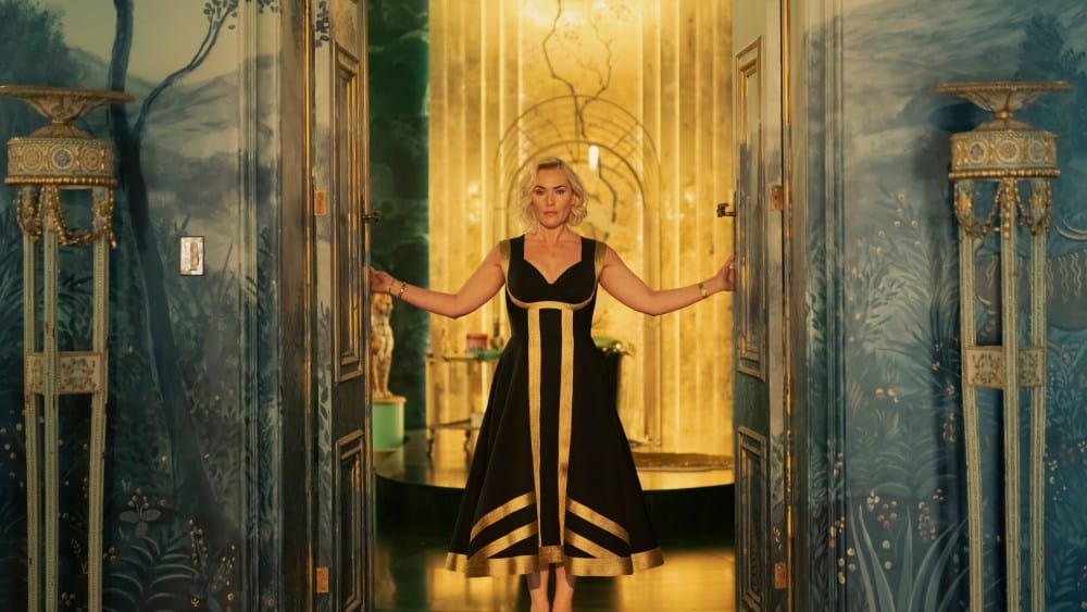 A woman resembling Kate Winslet, with blonde hair in a black and white dress, stands in a hallway with multiple doors, extending her arms out to touch the door handles. The walls are adorned with