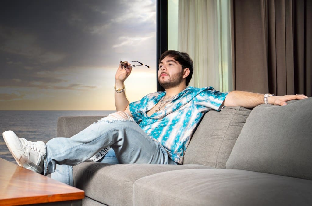 A young man lounges on a gray sofa, holding sunglasses, wearing a blue tie-dye shirt and distressed jeans, looking pensive. a scenic backdrop through a large window shows a sunset sky over the ocean.