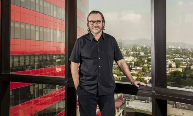A man in a black shirt and glasses stands on a balcony with one arm on the railing, smiling. Behind him are red building panels and a cityscape under a clear blue sky, reflecting the future