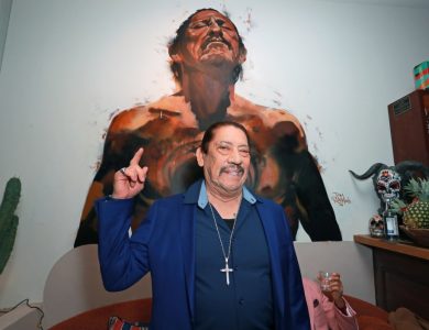 A Latino actor with a mustache, wearing a blue jacket and a cross necklace, smiles and points upwards. Behind him is a large painting of a man looking upwards with an expressive, brushy style