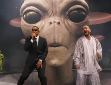 Two men perform on stage at Coachella, one in a black suit and sunglasses, the other in a shiny silver suit. Behind them is a large, realistic model of an alien head with big eyes