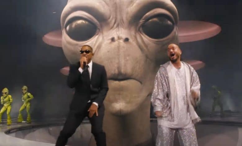 Two men perform on stage at Coachella, one in a black suit and sunglasses, the other in a shiny silver suit. Behind them is a large, realistic model of an alien head with big eyes