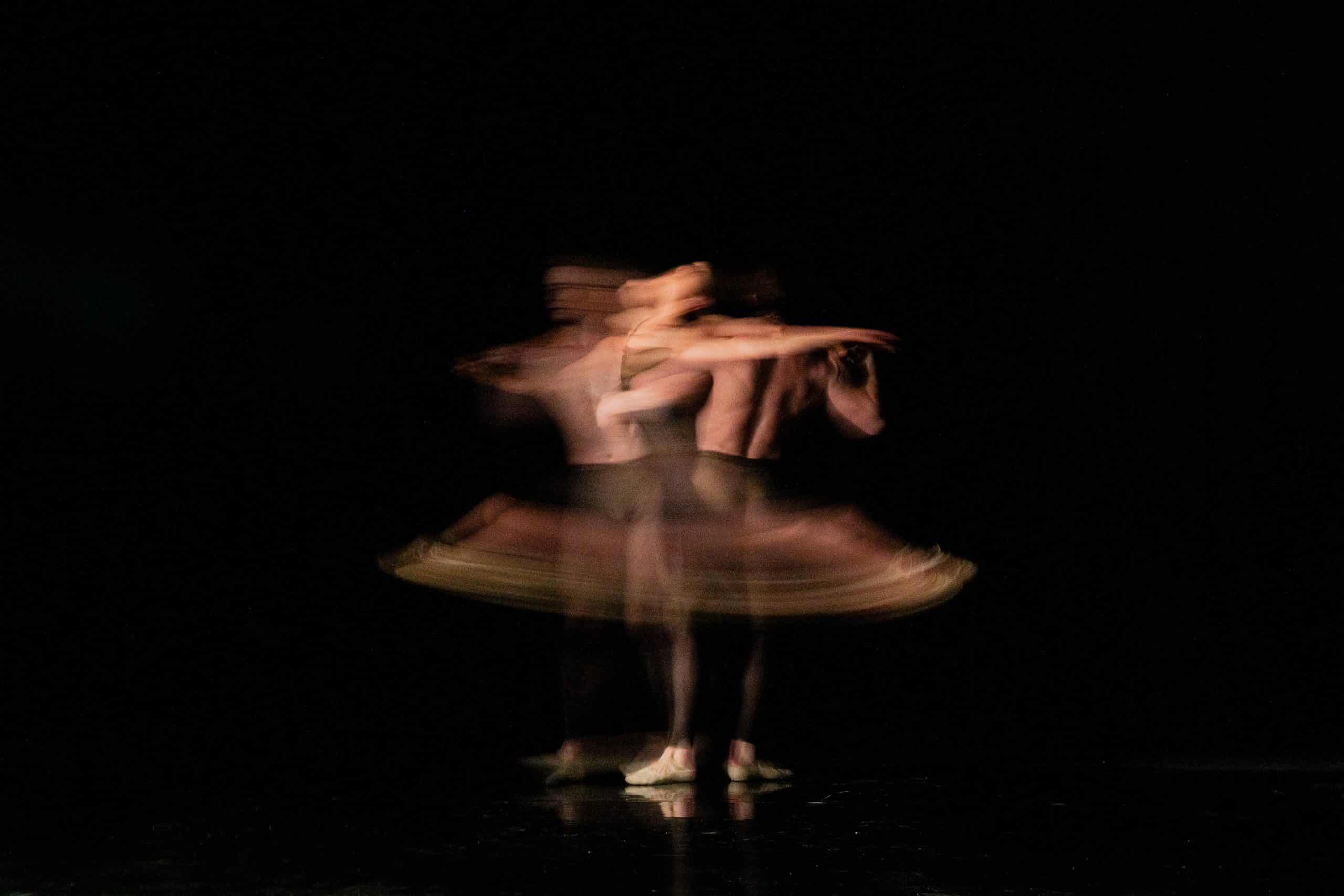 A blurred image of an American Ballet Theatre dancer in mid-twirl on a dark stage, capturing a sense of motion with multiple overlapping silhouettes suggesting rapid spinning.