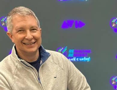 A smiling middle-aged man wearing a gray zip-up jacket, appointed as Avid's new CEO Wellford Dillard, sitting in front of a wall with a colorful pattern featuring repeating purple and green logos