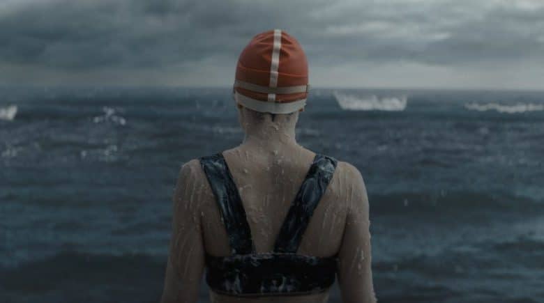 A young woman wearing a striped swim cap and a dark swimsuit stands facing the stormy sea, with her back to the camera. The sea is choppy under an overcast sky, exuding