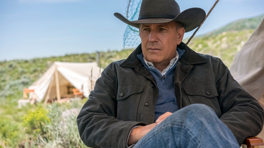 A man wearing a cowboy hat, navy jacket, and blue shirt sits outdoors, looking contemplatively to the side. In the background, there's a grassy field reminiscent of Yellowstone and a camp with