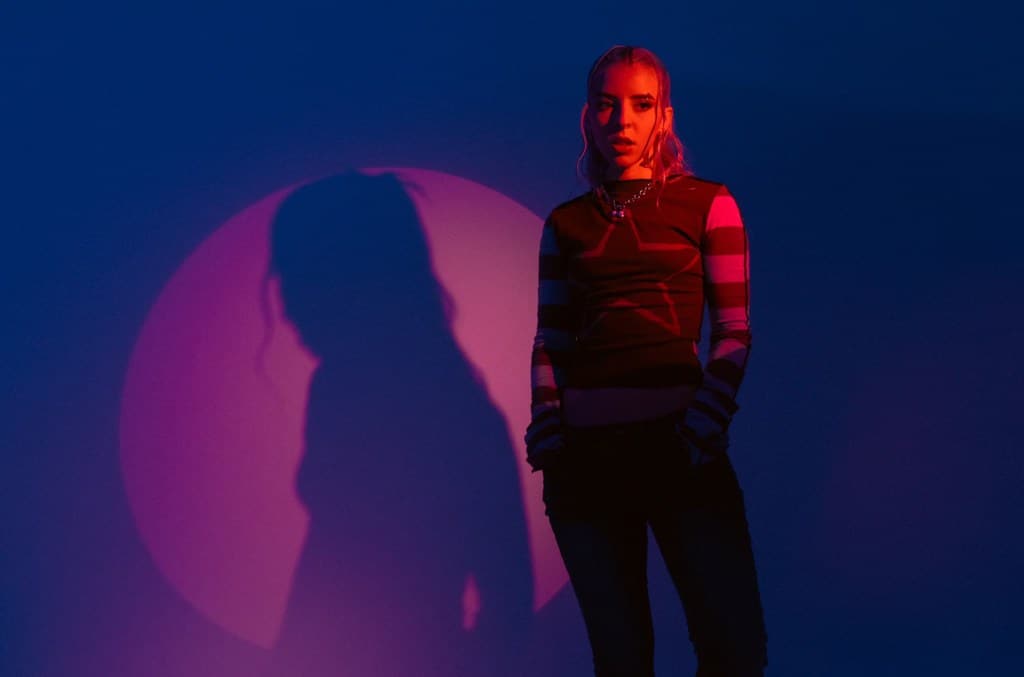 An Inside Look at Young Miko's Debut Album: A young artist stands in a moody, red-lit studio with a large circular shadow cast behind them. They wear a striped sweater and