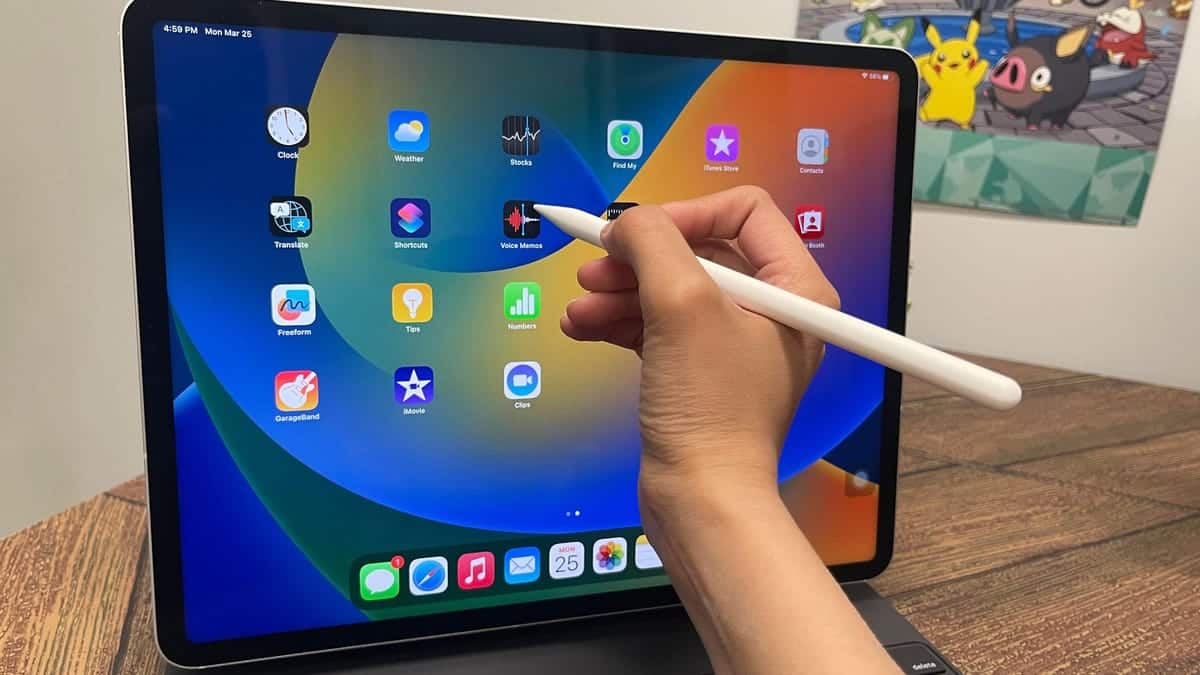 A person's hand holding a white Apple Pencil, about to interact with an iPad displaying a colorful app interface, with icons like Safari, Mail, and Photos. A Pikachu sticker is visible on the