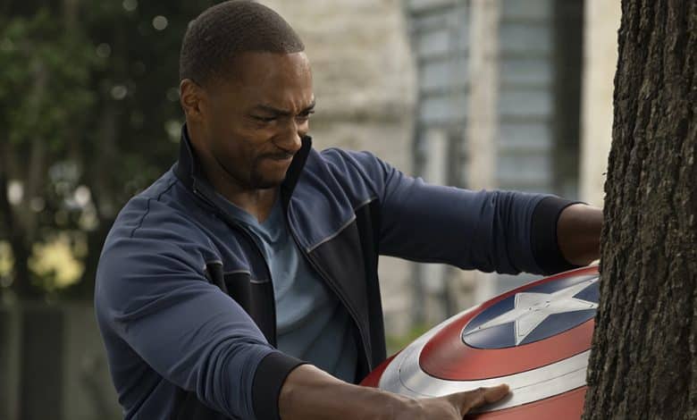 A man in a blue jacket strains as he grips a red, white, and blue shield pressed against a tree, showing determination on his face as Captain America.