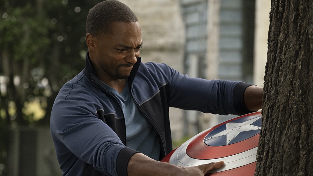 A man in a blue jacket strains as he grips a red, white, and blue shield pressed against a tree, showing determination on his face as Captain America.
