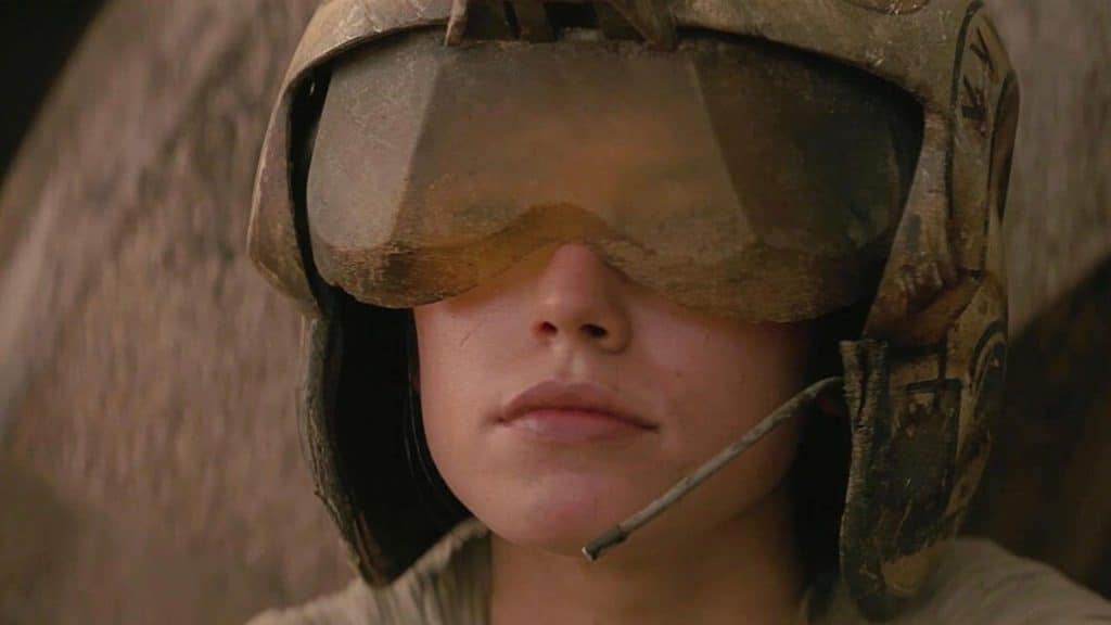 Close-up of a person wearing a military helmet and dusty goggles, partially obscuring their eyes. The helmet includes a mounted communication device. The background is blurred and indistinct, evoking scenes reminiscent