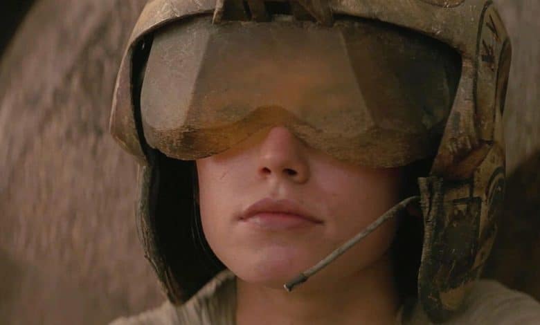 Close-up of a person wearing a military helmet and dusty goggles, partially obscuring their eyes. The helmet includes a mounted communication device. The background is blurred and indistinct, evoking scenes reminiscent