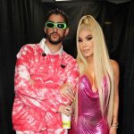 A man and a woman posing together, the man in a pink tie-dye hoodie and sunglasses, and the woman in a metallic pink dress with long blonde hair, standing in an area draped with black